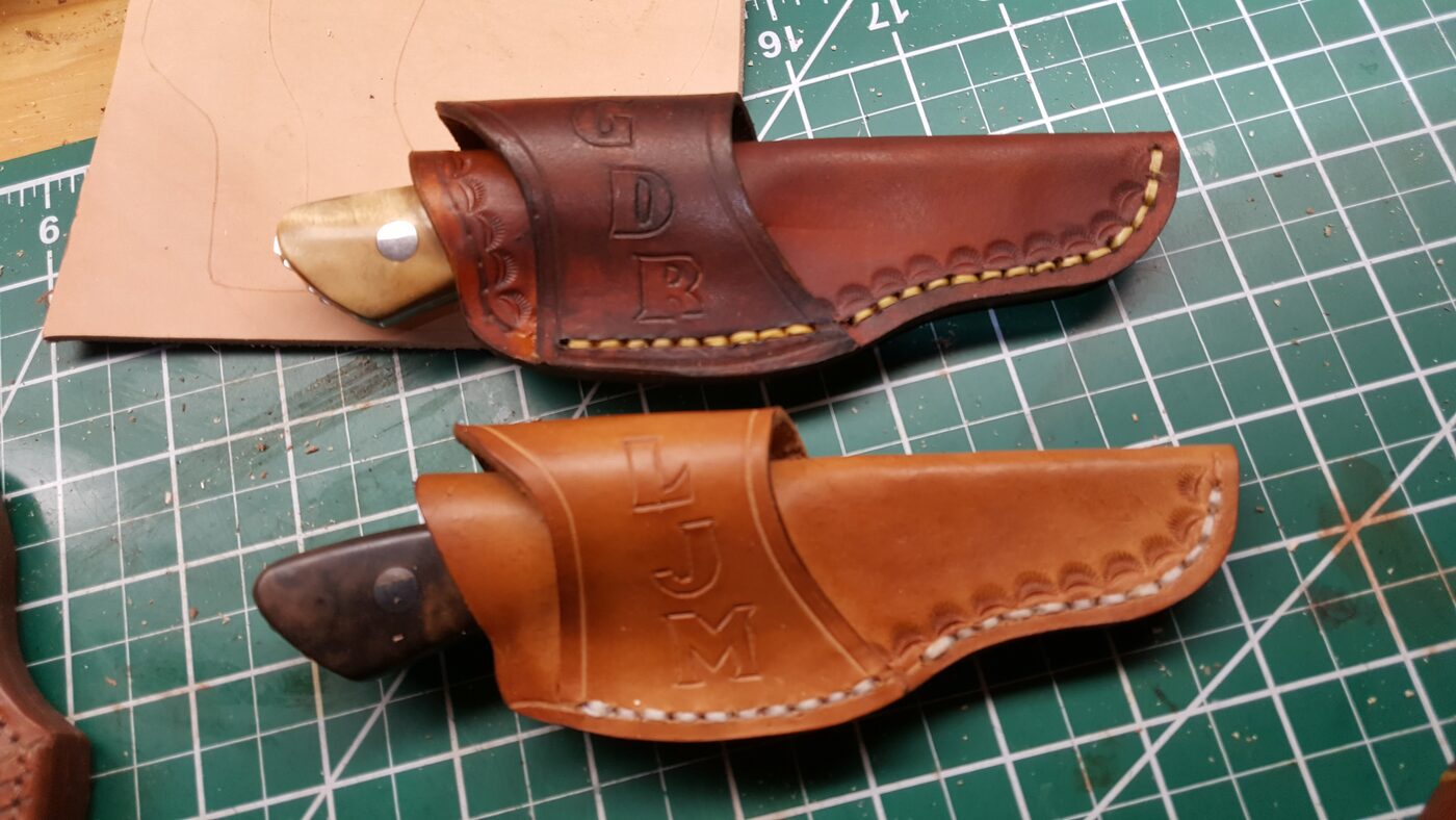 Two scout carry leather sheaths on a cutting mat, tooled with the owners' initials GDB and LJM, each fit to its own knife