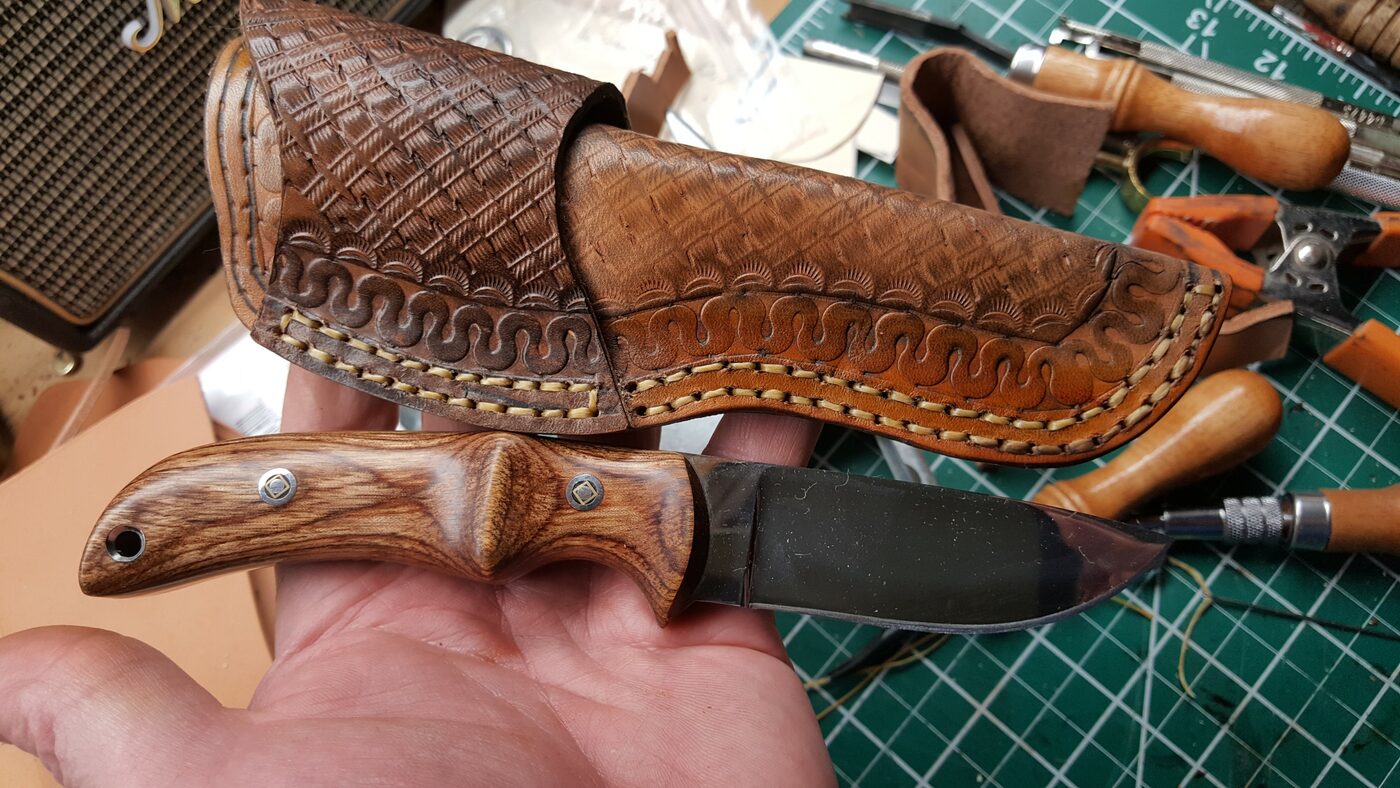 Plain natural russet leather scout carry sheath with a decorative stitched border, next to a wood-handled knife